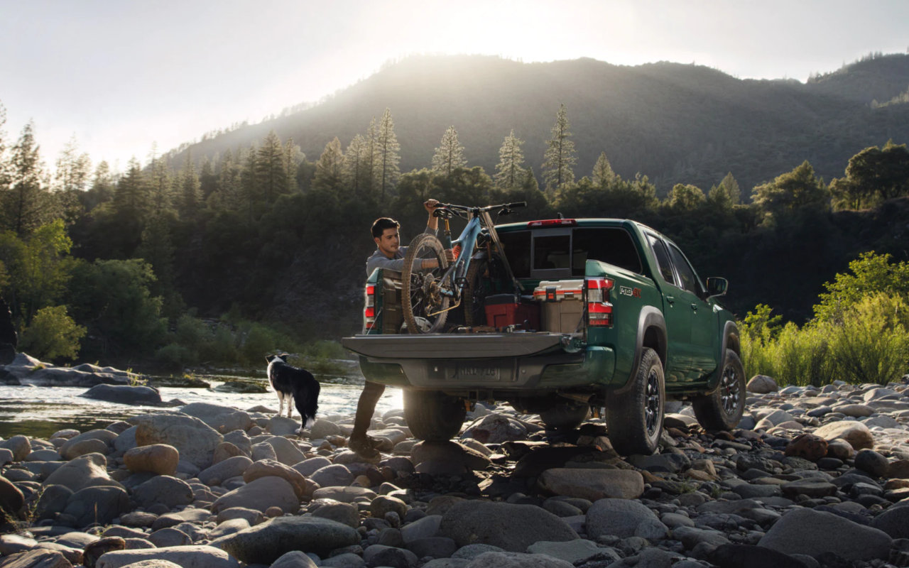 vue d'une Nissan Frontier 2023 sur le bord d'un cours d'eau avec son conducteur accompagné de son chien qui sort son vélo de montagne de la boite arrière